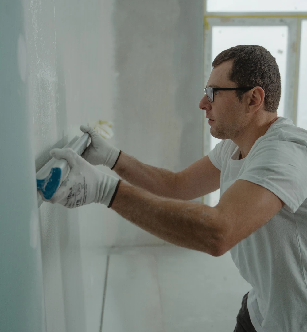 Man wearing glasses and gloves using a plaster smoothing trowel to level wet putty on a wall during interior renovation.