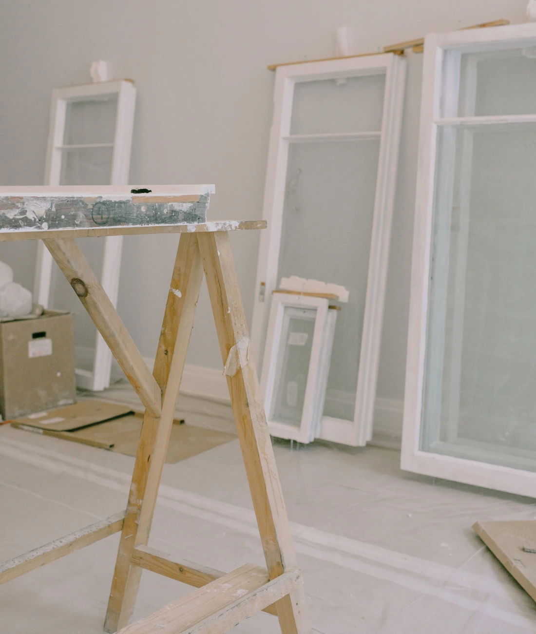 Wooden sawhorse standing in a brightly lit room under renovation, with white window frames resting against the wall in the background.